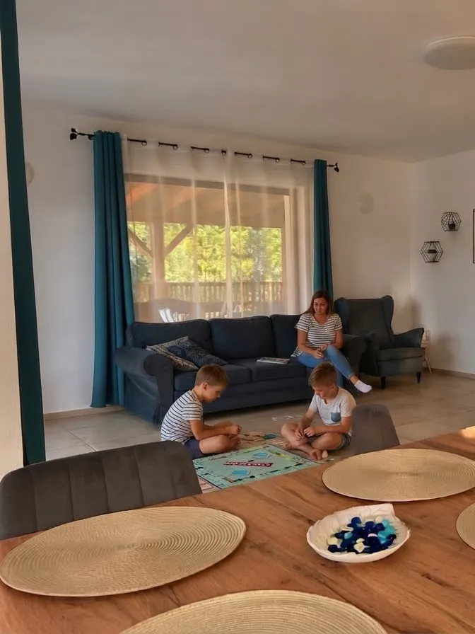 Livingul cu oaspeți: o femeie stă pe canapeaua albastră lângă fereastra cu perdele turcoaz, iar doi copii joacă un joc de societate pe covor, cu masa lungă în prim-plan
